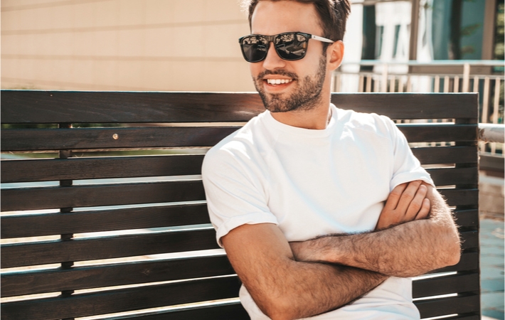 A smiling young adult man sitting on a bench with arms crossed and wearing a dark pair of sunglasses enjoying the sun outside.