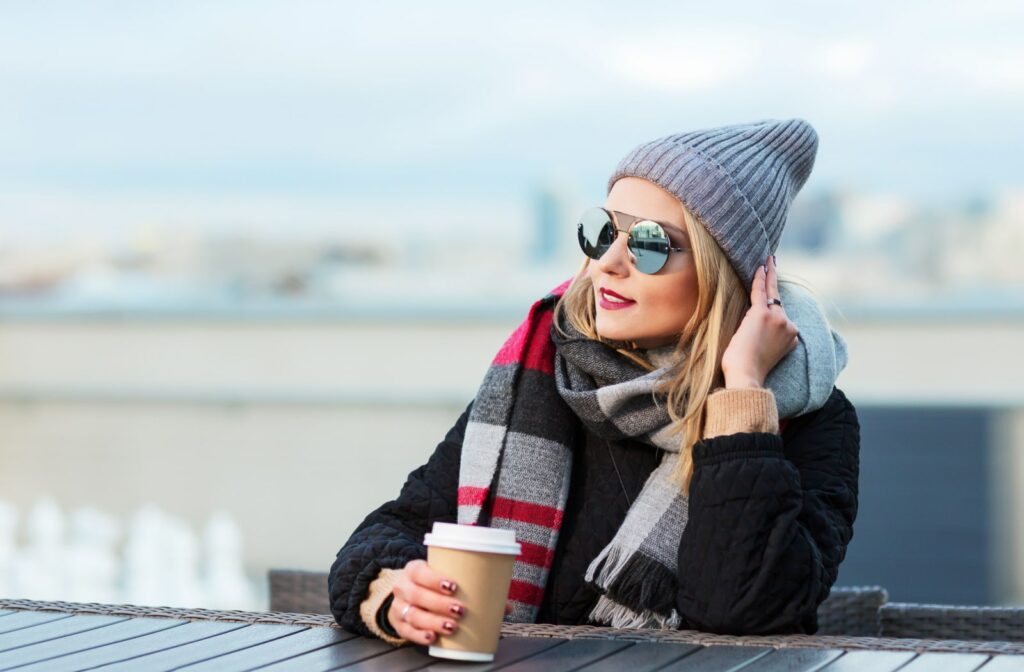 A woman sitting outside at a table and drinking a cup of coffee. She is wearing a winter jacket, hat, and sunglasses