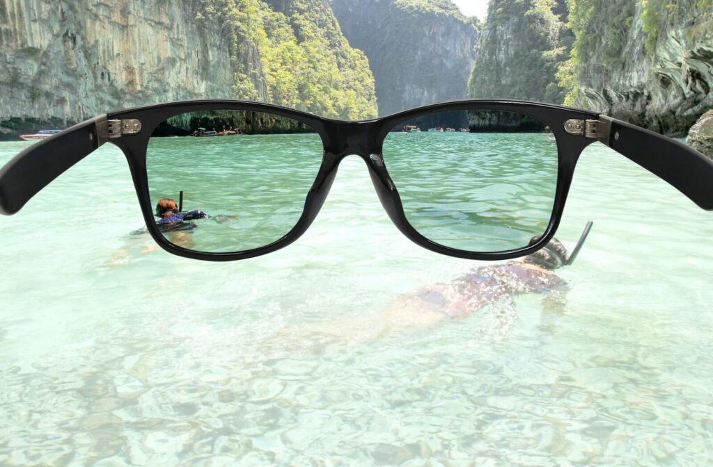 A photographer holds a pair of sunglasses up to the camera, while looking out at two people snorkelling in the turquoise waters of a beautiful tropical beach. The lenses remove the glare of the sun off the water, showing the lenses are polarized.