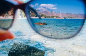 A person holds a pair of sunglasses up to the camera as they look out over a bay in a tropical island. The image through the glasses is clearly tinted, showing that the lenses are polarized.