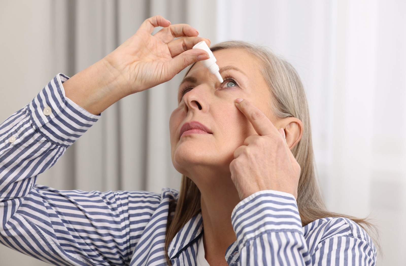 Middle-aged woman in striped shirt instilling eye drops in left eye.