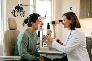 An optometrist examines a patient's eyes using a slit lamp in an exam room, with an eye chart on the wall.